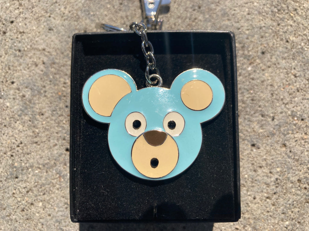 O. Bear blue and beige bear-shaped keychain on a black stand against a concrete background